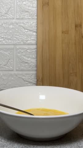 Vertical video close up. Man pours oatmeal from transparent plastic measuring cup into white bowl with beaten chicken egg yolks on kitchen table. Concept of cooking, breakfast, lunch, dinner, eat. Oat