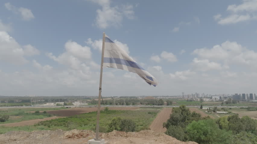 An Israeli flag on a mountain in the center of the country, with buildings and roads in the background under a blue sky with white clouds