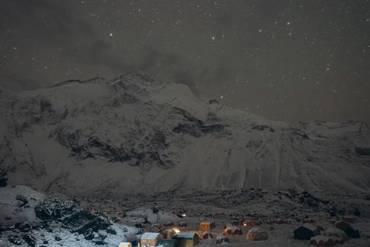 Starry night over a snow-covered campsite at the base of Mount Annapurna, Nepal, with glowing tents and a serene atmosphere, perfect for adventure enthusiasts.
