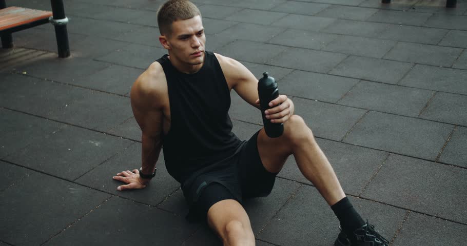 Athletic man taking break at workout, sitting on floor, resting, feeling thirsty and drinking water from bottle outdoors at sport ground. Healthy lifestyle concept. Strength, motivation. Outdoors gym.
