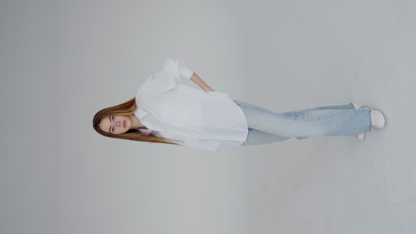 Studio portrait of a young woman dressed in a casual white shirt, demonstrating a simple yet striking pose against a plain background, highlighting modern minimal fashion. Young Woman. Vertical video.