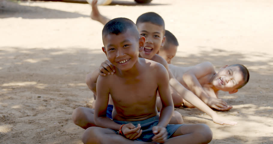 Children in a rural Thai village in Southeast Asia sit, smile, and laugh on wet ground under the shade of trees on a hot day, capturing the joy and simplicity of countryside life.