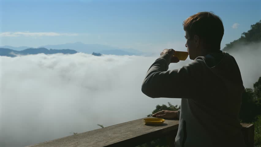 Man sitting on wooden deck while enjoying cup of coffee. Scenic view of mountains, clouds, and fog in background under bright blue sky. Serene and peaceful morning on high altitude spot
