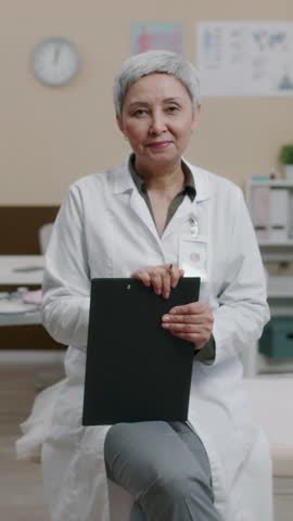 Vertical portrait of satisfied mature Caucasian female gynecologist with clipboard confidently posing for camera at workplace in hospital
