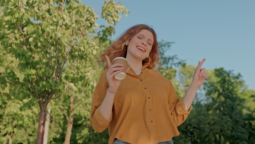 Joyfull woman dancing with coffee cup in hands. Cheerful female listening to music with wireless headphones. Red head woman relaxing at park after busy work day. Female having fun at city park.