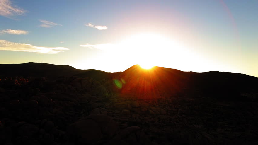 Aerial Backward Shot Of Sunrays Passing Over Dark Mountains In Desert During Sunset - Joshua Tree, California