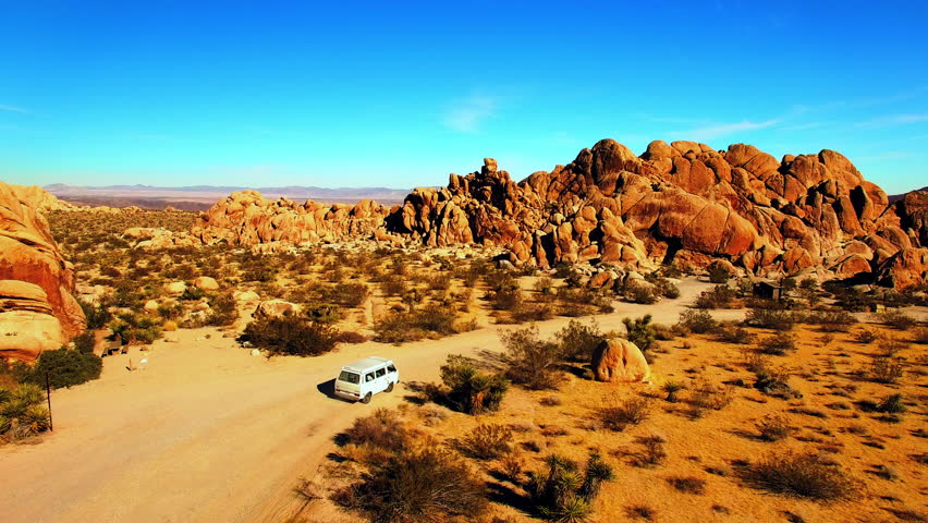 Aerial: Drone Panning Shot Of Off-Road Vehicle Moving In Semi Arid National Park Against Clear Sky On Sunny Day - Joshua Tree, California