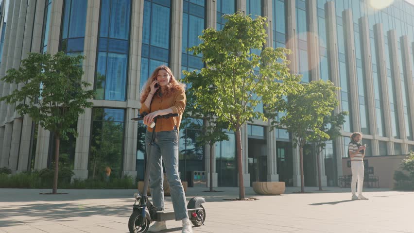 Stylish female conducting phone call outdoor. Woman leaned against electric scooter. Cheerful woman talking by gadget. Elegant red head female drinking coffee near modern glass building.