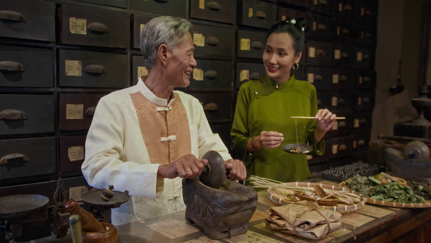 Medium shot of aged Asian male healer grinding ingredients and talking to female colleague at traditional medicine pharmacy