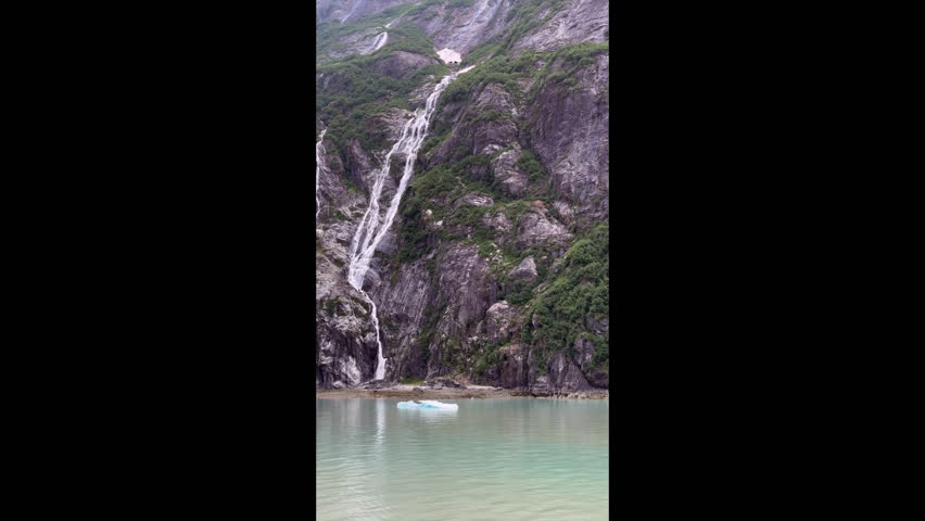 Narrow waterfalls down steep, glaciated cliff with spotty green vegetation along Tracy Arm fjord, with a solitary iceberg calved from North Sawyer Glacier, in southeastern Alaska. 4K vertical video.