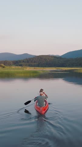 Active couple taking a canoe ride across the beautiful mountain lake, enjoying summer nature on a lakeshore vacation, aerial rear view shot. Vertical video