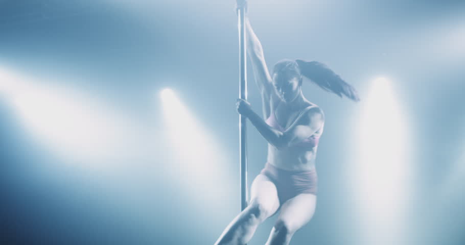 Slow-motion shot of a young woman pole dancing in dark interior with backlight and smoke