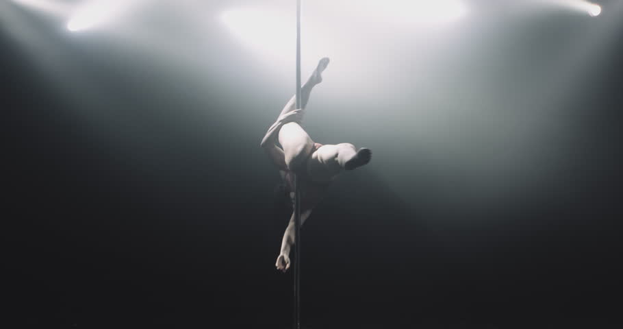 Dancer in dark studio spin on pole indoors. A silhouette looms against a smoky bright white background