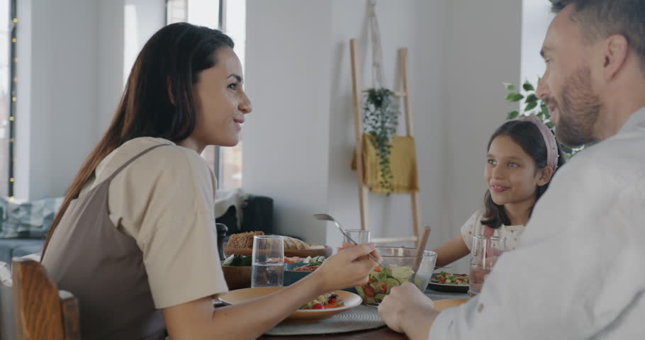Happy family mother father and daughter eating lunch and talking sitting at table in kitchen at home. Domestic meal and people concept.
