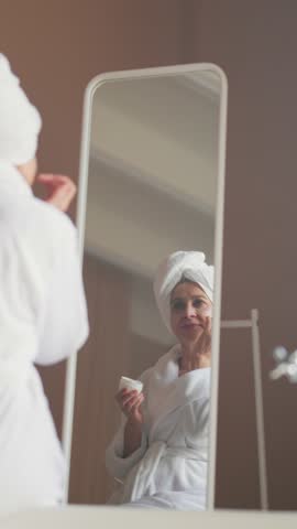 Skincare and daily morning routine of aged woman, vertical portrait of senior lady applying moisturizing cream on face. Old female person using cosmetic face mask after shower, anti-ageing face cream