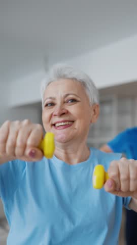 Positive old woman exercising with dumbbells at home, smiling and looking at camera, vertical video. Health care and sportive lifestyle in old age, stay active and healthy after 60 years, workout