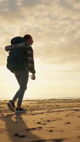 Woman backpacker walking alone on seacoast in summer day, carrying big backpack, travel in nature. Young adult female tourist traveling in beautiful places of planet, admiring and contemplating beauty