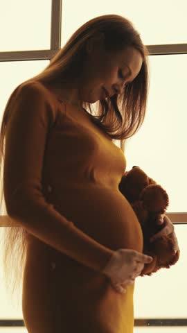Young adult pregnant woman stroking her belly and holding teddy bear, future mother, vertical video portrait. Beautiful and tender lady waits for baby, preparing to childbirth, eight months pregnancy