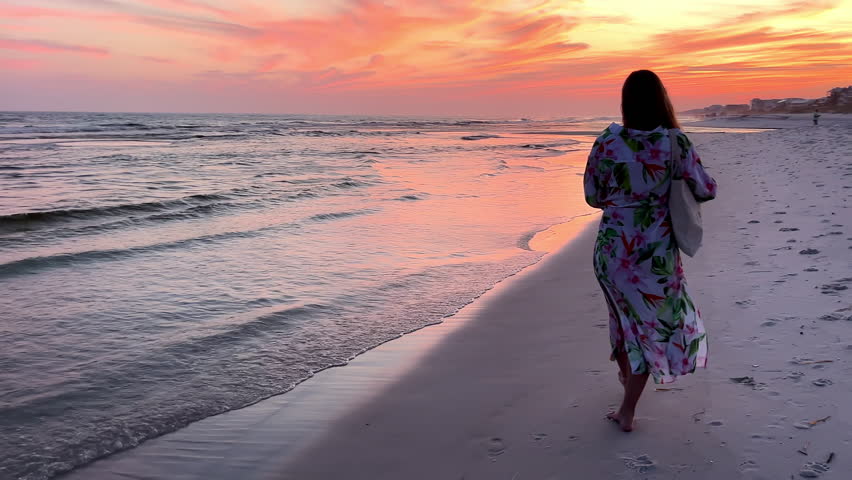 Woman Walking Along a Beach at Sunset, A woman walking along a serene beach during sunset, with the sky painted in vibrant colors.