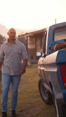 Vertical video portrait of smiling man unloading backpacks from pick up truck on road trip to cabin in countryside at sunset - shot in slow motion