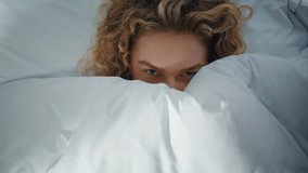 Relaxed girl lying bed hiding under blanket in morning. Closeup playful eyes peeking out cozy white bedding. Calm curly woman resting home enjoying weekend leisure. Beautiful blonde smile look camera. - Powered by Shutterstock - Get 15% off with code: PIKWIZARD15