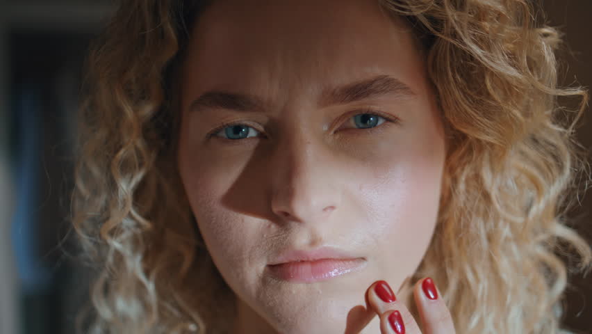 Pretty girl check pimple looking camera in sunlight closeup. Curly woman touch chin examining face skin problem in home mirror. Pensive blonde with acne. Dermatology skincare routine in morning.