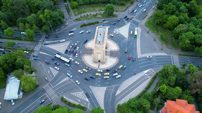 Aerial footage of the arc of triumph in Bucharest, Romania. High angle drone shot of boulevards, trees and a huge roundabout in europe on a summer afternoon. Gray asphalt and green parks from above.
