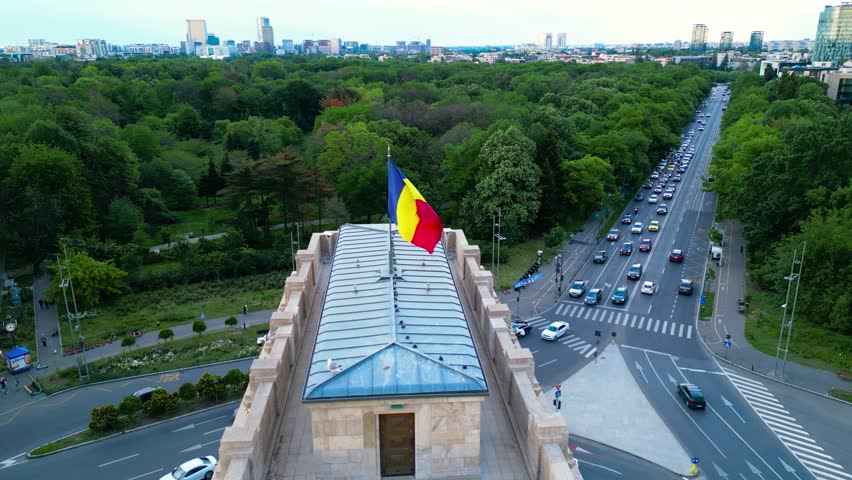 Aerial footage of the arc of triumph in Bucharest, Romania. High angle drone shot of boulevards, trees and a huge roundabout in europe on a summer afternoon. Gray asphalt and green parks from above.
