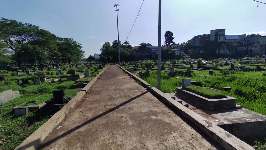 Long continuous take from Motorcycle POV entering the cemetery from a motor vehicle in the morning. Lots of tombstones. Public Burial place under trees on green grass in Jakarta, Indonesia. SSTKPlates