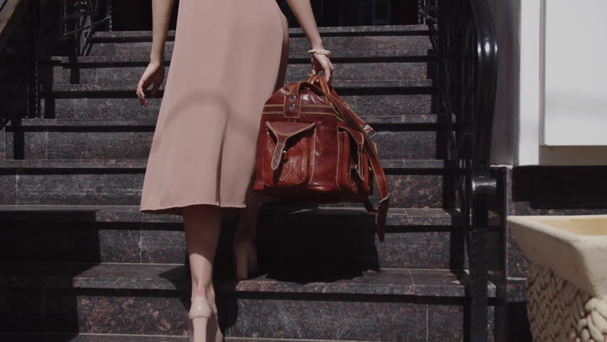 Girl in a dress with a leather travel bag is walking up the stairs, back view