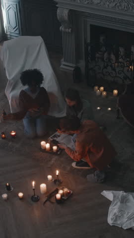 Vertical overhead shot of three teens sitting on floor among lit candles and reading incantations from occult book, invoking dark forces who live in dark desolate mansion