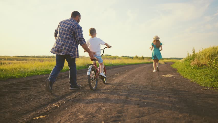 Happy family in summer vacation, parents teaching son to ride bike, rear view. Mother and father spending time with child in nature, parents running near little boy riding bicycle, following shot