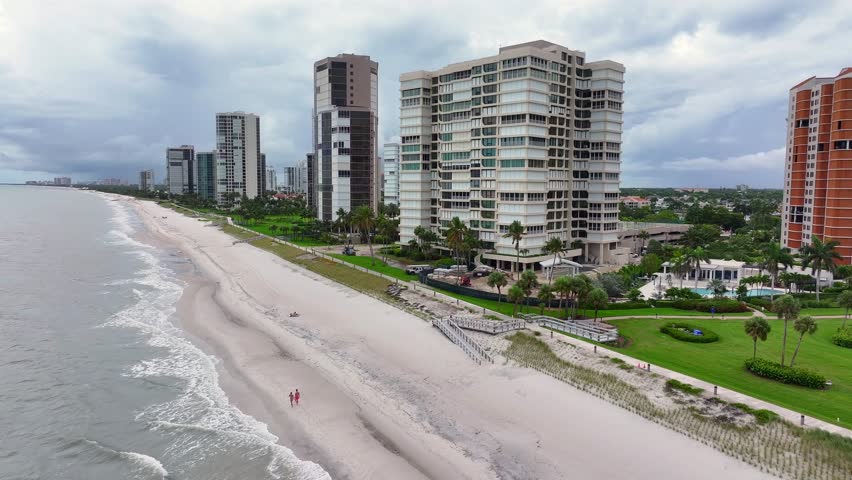 Downtown Apartments along coastline of Naples with sandy beach and walking people. Florida, USA. Cloudy day over Rising drone wide shot.