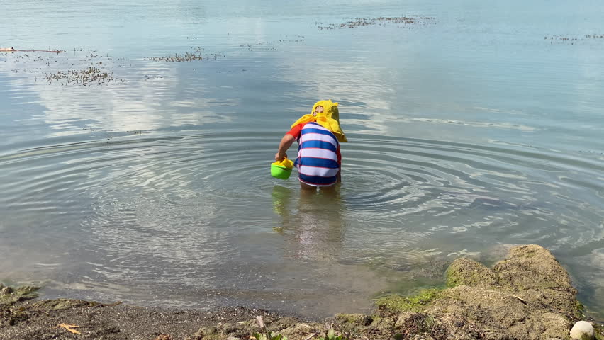 Small child playing on the lake shore on a summer day. Little boy wearing sun protection shirt in nature. Simferopol, Crimea, Russia 2024.