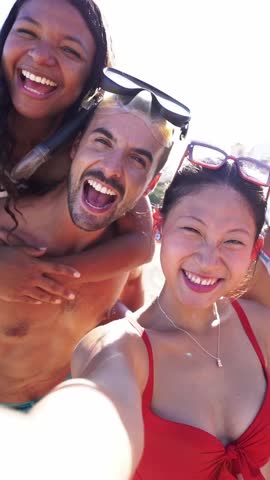 Vertical selfie video-call of multiethnic of friends on the beach having fun outdoors looking at the camera smiling. Young multiethnic people in swimsuit enjoying summer holidays.