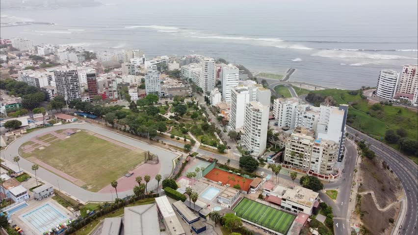 Aerial view of coastal city with high-rise buildings, parks, and scenic shoreline. Roads winding through lush green areas