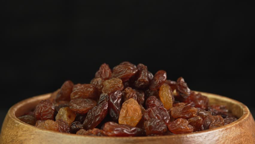 Black raisins slowly rotating in wooden bowl on black background. Dried grapes close up. Dried fruits and dry berries