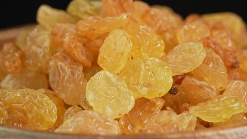 Green raisins slowly rotating in wooden bowl in macro on black background. Dried fruits and dry berries. Dried grapes close up