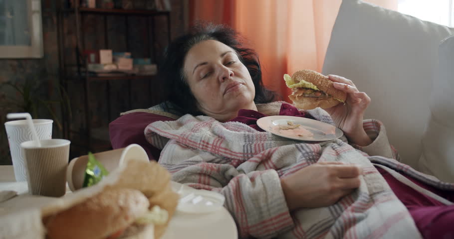Unkept lazy woman lying on the couch, eating fast food. Wants to reach for a soda, but lazily refuses the idea. Messy used fast food trash on the table at home. Careless, laziness concept. 