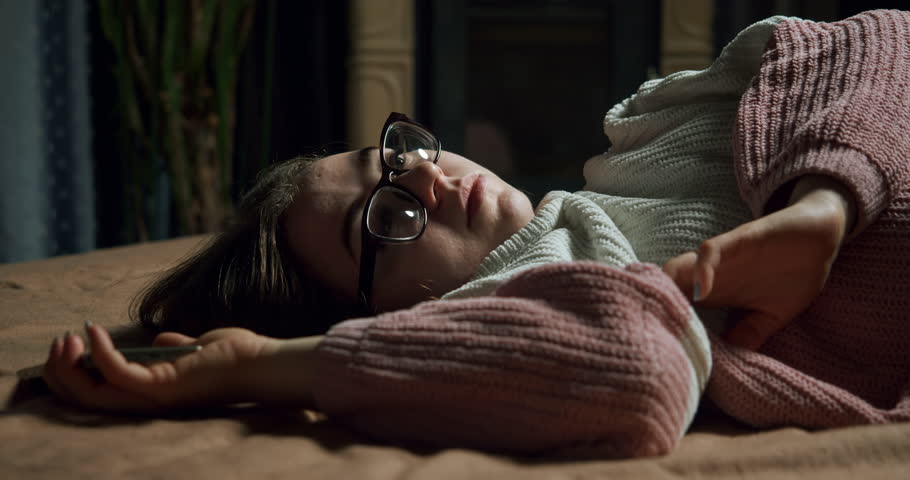 Bored teen girl in glasses using smartphone, wait for important message, interesting information update. Relationship problem. Move phone away with upset. Lying on bed in a sad bad mood