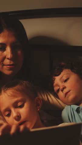 Vertical screen: Mother and her two children cuddle in bed while daughter reads bedtime story. Concept of family bonding and bedtime rituals. Focus on love, comfort, and nurturing environment