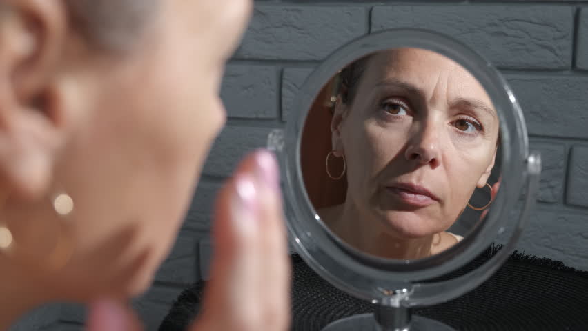 Suffer from face age related changes. A view of depressed and frustrated look at her aged changes on her face in the mirror.
