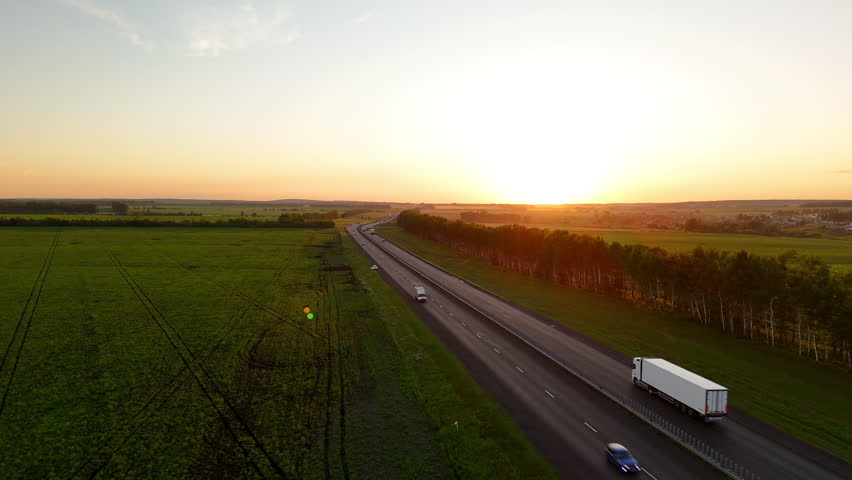 Aerial view of the highway in the rays of the setting sun and a moving truck with a semi-trailer. Heavy truck driving on an asphalt road along green fields. Cargo delivery, transportation.