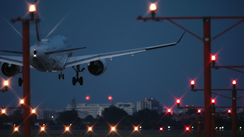 Rear view passenger airplane landing on runway in slow motion. Close-up unrecognizable white aircraft lands on airport runway at night. Signal pylon lights foreground. Heat distortions effect
