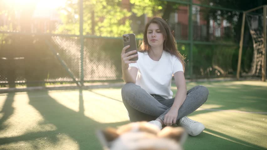 A young, beautiful female blogger in a white T-shirt and jeans is broadcasting live from her phone. The girl is streaming while her fluffy corgi is playing around her