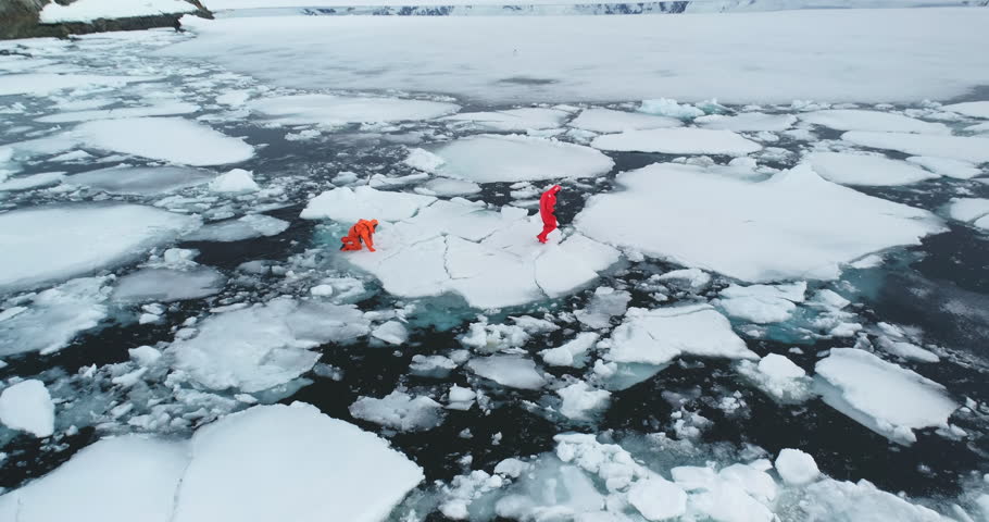People in orange immersion suits run melting ice, Antarctica. Men in wetsuits try to escape from polar ocean trap, fall in cold water ice floes. Explore climate, global warming, melt icebergs. Aerial
