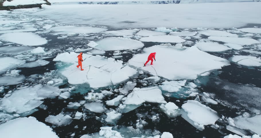 People in orange immersion suits run melting ice, Antarctica. Men in wetsuits try to escape from polar ocean trap, fall in cold water ice floes. Explore climate, global warming, melt icebergs. Aerial