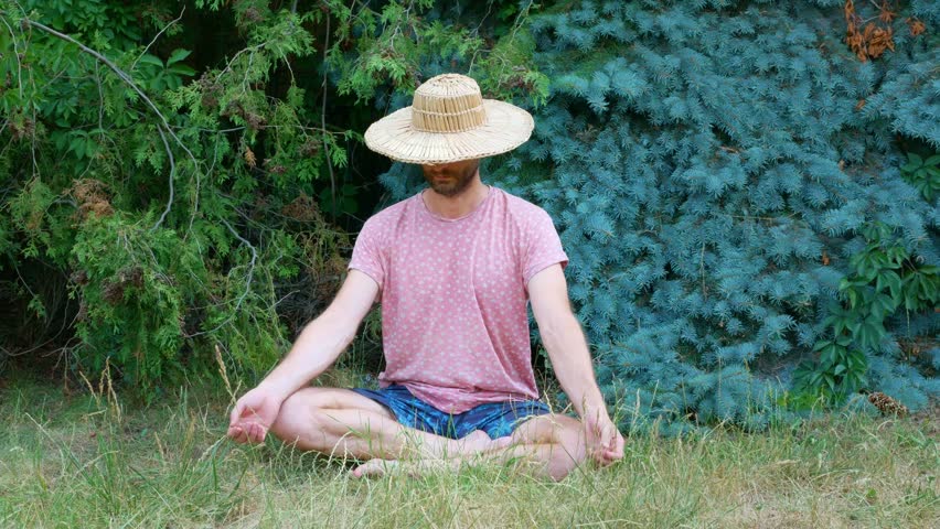 A man in a straw hat with a beard meditates in a summer park