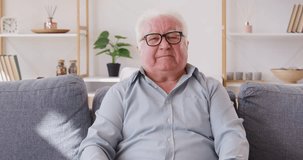 Smiling elderly man sitting on a couch, looking at the webcam, and waving hand in greeting while talking online. He appears happy and engaged in the internet video call communication from home. - Powered by Shutterstock - Get 15% off with code: PIKWIZARD15