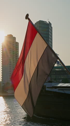 Vertical video. Real large Netherlands Flag waving in de wind at the cityscape at sunset. The Dutch flag is a national symbol of the Netherlands. Dutch red white blue flag